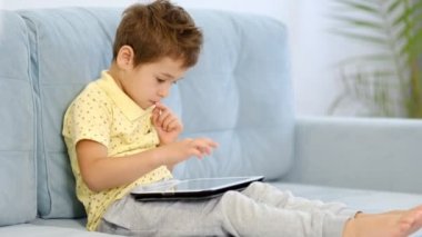 Anaokulu çocuğu evdeki kanepede otururken online eğitim için tablet bilgisayar kullanıyor. Çocuk internetten okumak için başvuru kullanıyor. Aleti olan çocuk.