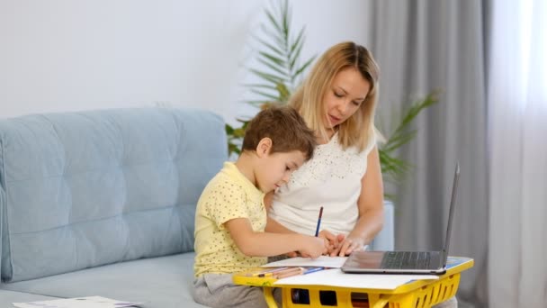 Junge Mutter oder Kindermädchen hilft einem kleinen Jungen eine Aufgabe in einer Online-Schule zu erledigen. Mutter und Sohn machen gemeinsam Hausaufgaben in der Schule — Stockvideo