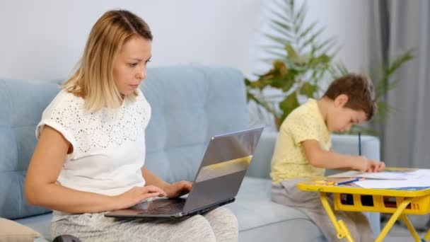 Maman souriante travaillant à la maison avec son enfant sur le canapé tout en écrivant un e-mail. Jeune femme travaillant à domicile, en quarantaine. Calme jeune mère ou nounou assis sur le canapé travaillant sur ordinateur portable — Video