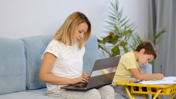 Maman souriante travaillant à la maison avec son enfant sur le canapé tout en écrivant un e-mail. Jeune femme travaillant à domicile, en quarantaine. Calme jeune mère ou nounou assis sur le canapé travaillant sur ordinateur portable — Video