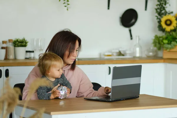 Mom working remotely on laptop while taking care of her baby. Young mother on maternity leave trying to freelance by the desk with toddler child. Close up, copy space, background. — Stock Photo, Image