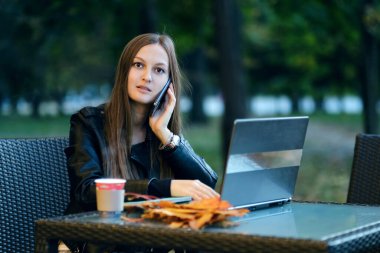 Genç ve güzel bir iş kadını sokak kafesinde dizüstü bilgisayarda çalışıyor, klavyede daktilo kullanıyor, akıllı bayan cidden ekrandaki akıllı telefona ve masadaki gözlüğe bakıyor. Şık pembe ceketli, beyaz saatli..