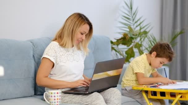 Maman souriante travaillant à la maison avec son enfant sur le canapé tout en écrivant un e-mail. Jeune femme travaillant à domicile, en quarantaine. Calme jeune mère ou nounou assis sur le canapé travaillant sur ordinateur portable — Video
