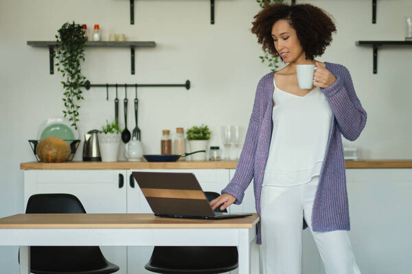 Happy woman holding a cup of coffee and checks mail on his laptop in her kitchen. Young african american female drinking coffee in her kitchen. relaxed morning concept