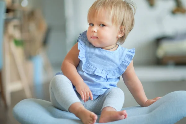 Porträtt av en blond blåögd flicka 1 år gammal sitter på en blå stol. Söt liten bebis leker hemma i rummet. — Stockfoto