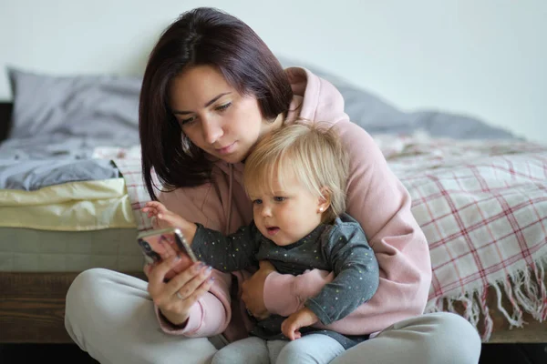 A kislány ül az anyjával az ágyon, és néz az okostelefon rajzfilmek, játszik játék. A gyerek videón keresztül beszél a nagyanyjával. Anya képeket mutat a lányának a képernyőn.. — Stock Fotó