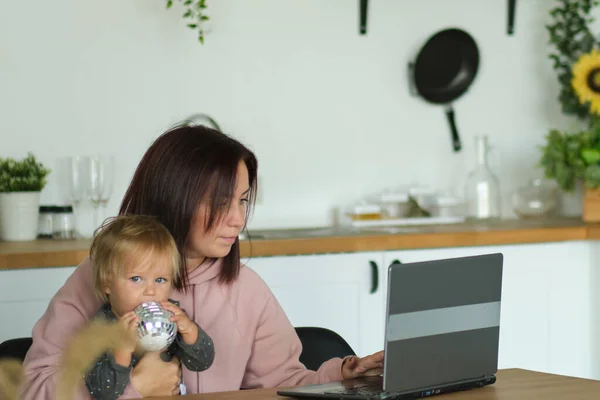 Mom working remotely on laptop while taking care of her baby. Young mother on maternity leave trying to freelance by the desk with toddler child. Close up, copy space, background. — Stock Photo, Image