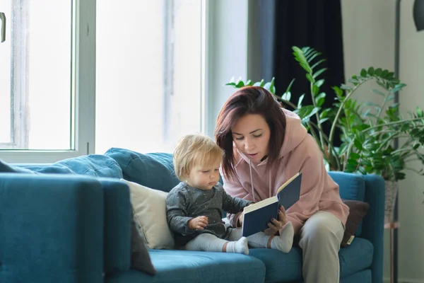 Anya könyvet olvas a babának a kanapén. lefekvés előtt. Vigyázok a gyerekekre. Az anyák szeretik a lányukat. — Stock Fotó