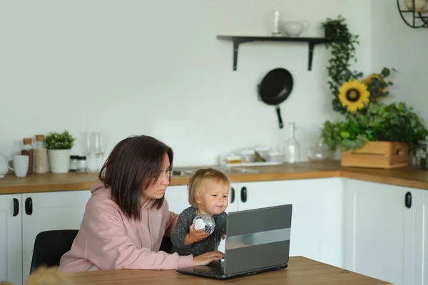 Anya távolról dolgozik a laptopon, miközben vigyáz a babájára. Fiatal anya szülési szabadságon próbál szabadúszó lenni az asztalnál egy kisgyerekkel. Közelkép, másolás tér, háttér. — Stock Fotó