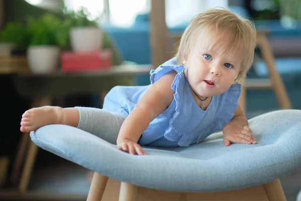 Little baby tries to climb on the chair on her own. Cute little baby playing at home in room. — Stock Photo, Image