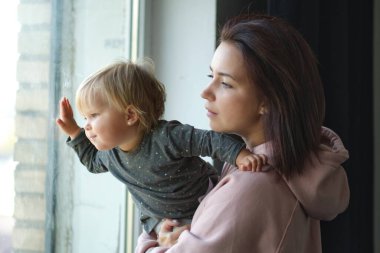 Şefkatli anne ve bebek birlikte pencerenin yanında, Otantik aile. Sevimli, tombul, beyaz bir kız, annesi pencere eşiğinde ve pencereden dışarı bakıyor..