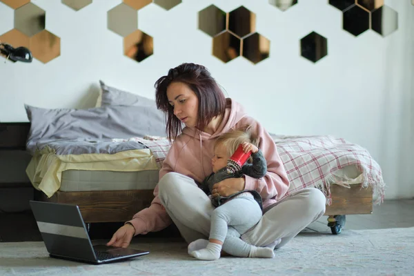 A fiatal anya a gyerekével a földön ül és egy laptopon próbál dolgozni. diktatúrában dolgozni. online tanulás fiatal anyák számára. szakmát szerezni otthonról távolról. — Stock Fotó