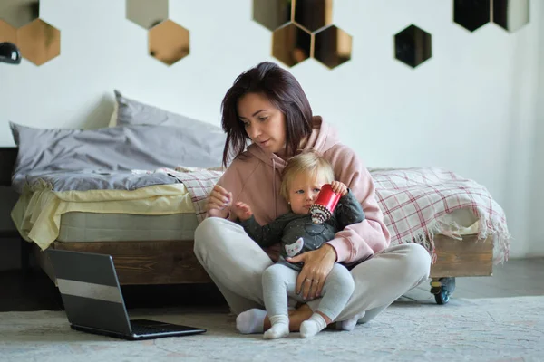 Fiatal, elfoglalt anya az imádnivaló kislányával, aki laptopon dolgozik vagy tanul. Otthon dolgozó nő babával az ölében — Stock Fotó