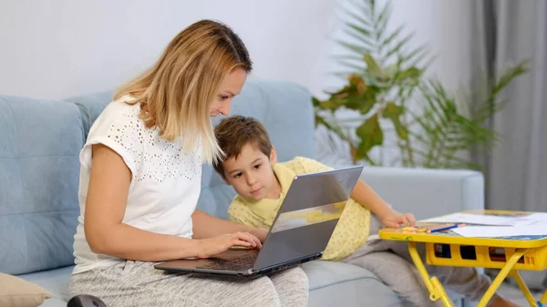 在写电子邮件的时候，笑着妈妈和她的孩子在沙发上干活。年轻妇女在家里工作，被隔离在隔离环境中。年轻妈妈或保姆安静地坐在沙发上，在笔记本电脑上工作 — 图库照片
