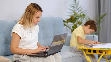 Gülümseyen anne, e-posta yazarken çocuğuyla birlikte koltukta çalışıyor. Karantina karantinasında çalışan genç bir kadın. Sakin ol genç anne ya da dadı kanepeye otur ve dizüstü bilgisayarla çalış.