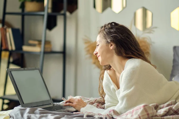 Boldog alkalmi gyönyörű nő dolgozik egy laptop ül az ágyon a házban. — Stock Fotó