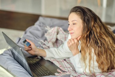 Kahverengi saçlı neşeli bir kadın internette laptopuyla yatak odasında alışveriş yapıyor.