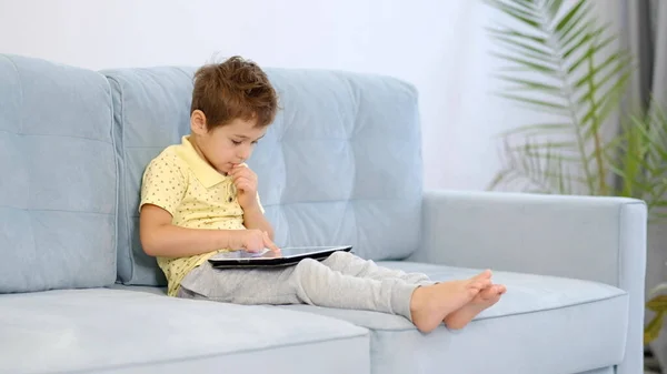 Αγόρι σπίτι κάθεται στον καναπέ να παίξει φαίνεται δισκίο κινούμενα σχέδια παιχνίδια ταινία — Φωτογραφία Αρχείου