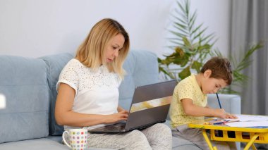 Gülümseyen anne, e-posta yazarken çocuğuyla birlikte koltukta çalışıyor. Karantina karantinasında çalışan genç bir kadın. Sakin ol genç anne ya da dadı kanepeye otur ve dizüstü bilgisayarla çalış.