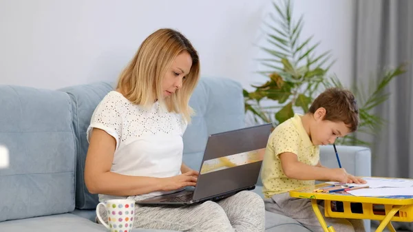 Leende mamma arbetar hemma med sitt barn på soffan medan du skriver ett mail. Ung kvinna som arbetar hemifrån, i karantän isolering. Lugn ung mor eller barnflicka sitta på soffan arbetar på laptop — Stockfoto