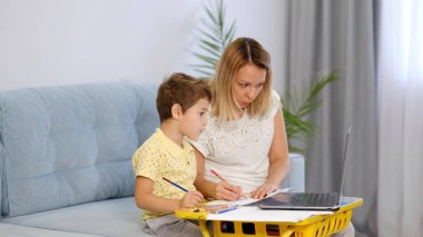 Genç bir anne ya da dadı, küçük bir çocuğun online bir okulda ödevini tamamlamasına yardım eder. Anne ve oğul birlikte okul ödevi yapıyorlar.
