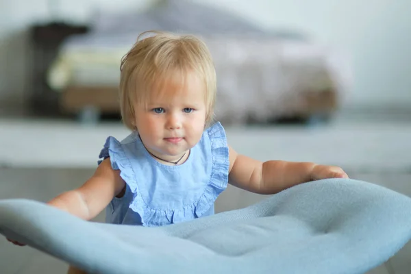 Маленька блакитноока дівчинка вчиться ходити, тримаючись за стілець. щасливе дитяче обличчя — стокове фото