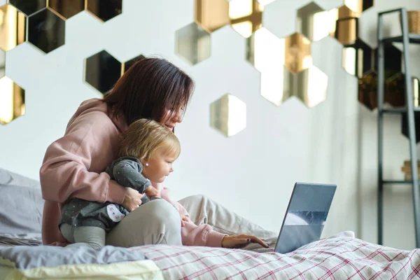 Anya távolról dolgozik a laptopon, miközben vigyáz a babájára. Fiatal anya szülési szabadságon próbál szabadúszó lenni az asztalnál egy kisgyerekkel. Közelkép, másolás tér, háttér. — Stock Fotó