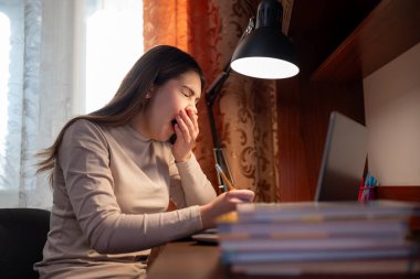 Ev ödevi sırasında yorgunluktan esneyen bir kız öğrenci. Genç bir kız dizüstü bilgisayarla masada oturuyor ve çalışmaktan yorulduğunu gösteriyor.