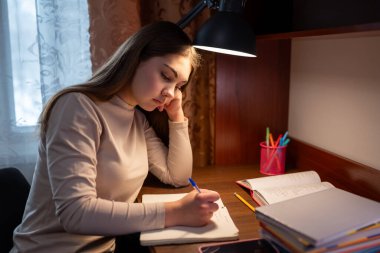 Evde sınavlara hazırlanan yorgun bir kız öğrenci. Genç kız, yorgun ve üzgün, masada oturuyor ve ödev yapıyor, evde eğitim görüyor.