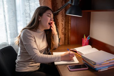 Çalışırken yorgunluktan esneyen bir kız öğrenci. Ders çalışmaktan bitap düşmüş genç bir kız bir masada oturuyor ve eliyle ağzını kapatıyor.
