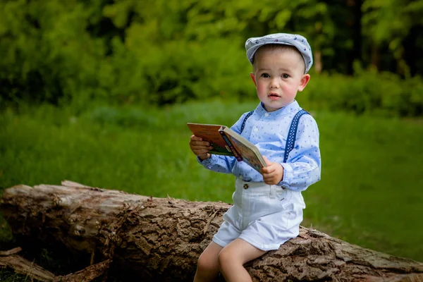 Ein kleiner zweijähriger Junge sitzt auf einem Baum und liest ein Buch. Kindertag — Stockfoto