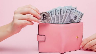 Üzerinde dolar olan pembe cüzdan ve fotokopi alanı olan pembe arka planda kadının elinde bitcoin..