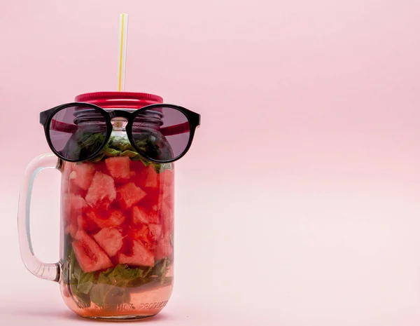Glasses lying on a Jar of cold fresh lemonade with piece of watermelon and ice on pink background — Stock Photo, Image