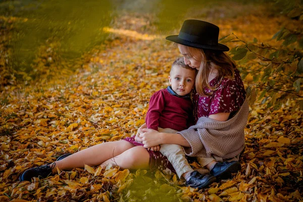 Счастливая молодая мать, играющая с ребенком в осеннем парке с желтыми лопастями. Маленький мальчик с матерью играют в парке осенью — стоковое фото