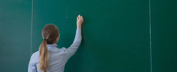 Pandangan belakang seorang siswa atau guru dengan rambut cokelat panjang menulis di papan tulis hijau kosong atau papan tulis dengan ruang penyalinan — Stok Foto