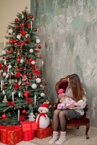 Merry Christmas and Happy Holidays Pretty young mom reading a book to her cute daughter near Christmas tree indoors.