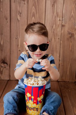 Küçük şirin çocuk bebek çocuk 2-3 yaşında, patlamış mısır, ahşap arka plan üzerinde hızlı yemek yeme için kova tutarak 3d IMAX sinema gözlük. Çocuklar çocukluk yaşam tarzı kavramı. Kopya alanı.