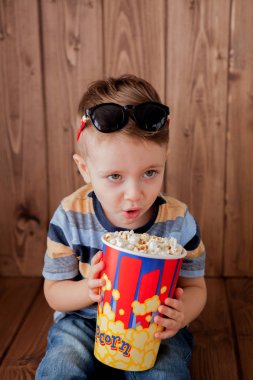 Küçük şirin çocuk bebek çocuk 2-3 yaşında, patlamış mısır, ahşap arka plan üzerinde hızlı yemek yeme için kova tutarak 3d IMAX sinema gözlük. Çocuklar çocukluk yaşam tarzı kavramı. Kopya alanı.