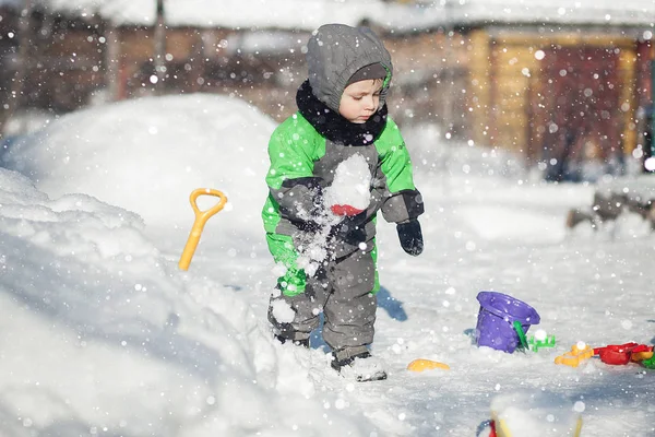 Porträt eines niedlichen Kleinkindes, das auf Schnee sitzt und mit seinem gelben Traktorspielzeug im Park spielt. Kind spielt im Freien. Glücklicher Junge mit Bauspielzeug. Lifestylekonzept — Stockfoto