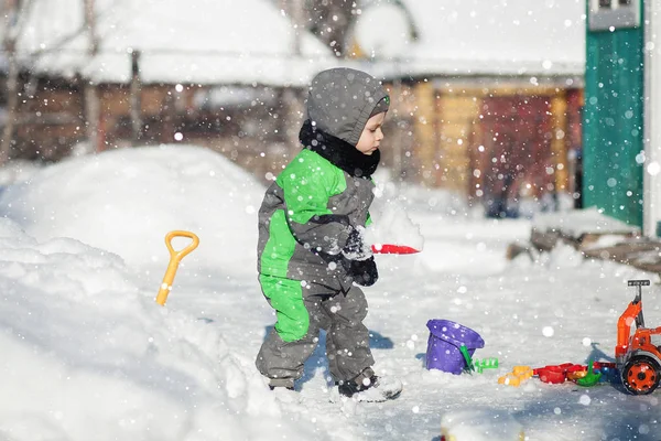 Ritratto di piccolo bambino carino seduto sulla neve e giocare con il suo giocattolo trattore giallo nel parco. Bambino che gioca all'aperto. Ragazzo felice con giocattolo da costruzione. Stile di vita concetto — Foto Stock