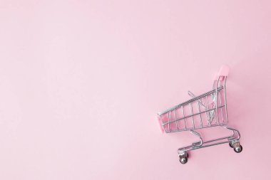 Küçük süpermarket bakkal itme sepeti oyuncak alışveriş için tekerlekler ve pembe plastik elemanları pembe pastel renkli kağıda düz arka plan yattı. Alışveriş kavramı. Kopya alanı reklam için.