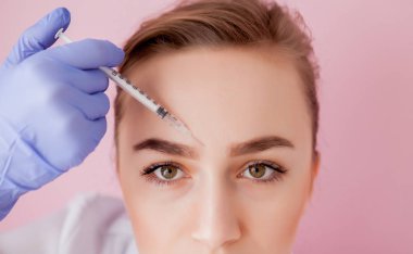 Doktor kozmetik uzmanı güzellik salonundaki genç ve güzel bir kadının cildindeki kırışıklıkları sıkmak ve düzleştirmek için yüz enjeksiyonlarını yenileme prosedürünü uyguluyor..
