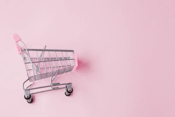Small supermarket grocery push cart for shopping toy with wheels and pink plastic elements on pink pastel color paper flat lay background. Concept of shopping. Copy space for advertisement.