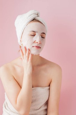 Güzel genç kadın kozmetik doku maske pembe bir arka plan üzerinde bir yüzünde uyguluyor.
