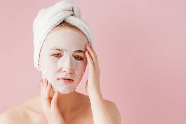 Güzel genç kadın kozmetik doku maske pembe bir arka plan üzerinde bir yüzünde uyguluyor.