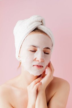 Güzel genç kadın kozmetik doku maske pembe bir arka plan üzerinde bir yüzünde uyguluyor.