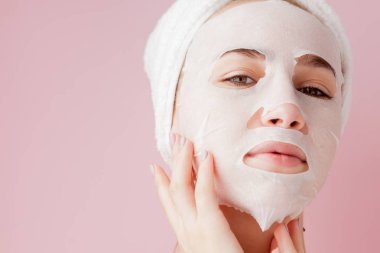 Güzel genç kadın kozmetik doku maske pembe bir arka plan üzerinde bir yüzünde uyguluyor.