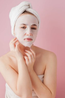 Güzel genç kadın kozmetik doku maske pembe bir arka plan üzerinde bir yüzünde uyguluyor.
