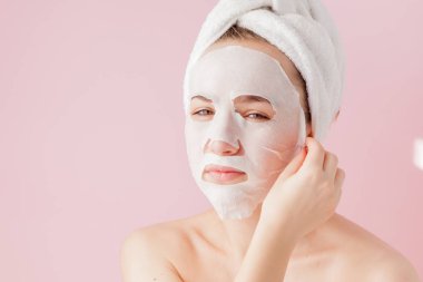 Güzel genç kadın kozmetik doku maske pembe bir arka plan üzerinde bir yüzünde uyguluyor.
