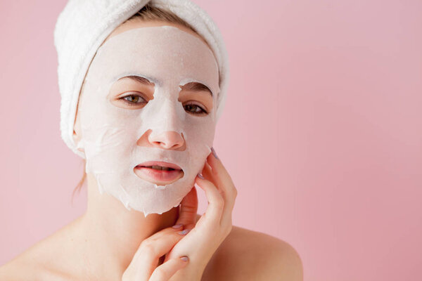Beautiful young woman is applying a cosmetic tissue mask on a face on a pink background.